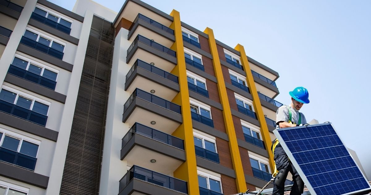 Energia Solar Vantagens e Desvantagens: Energia solar em varandas de apartamentos - Um trabalhador de capacete azul instala um painel solar em um telhado com o fundo de um edifício de apartamentos moderno, destacando a integração de energias renováveis em ambientes urbanos.