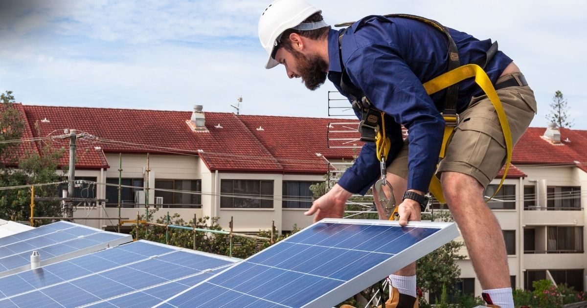 Energia Solar Vantagens e Desvantagens - Trabalhador com capacete branco e equipamento de segurança ajusta painéis solares no telhado, com casas residenciais ao fundo, demonstrando a expansão da energia solar em áreas residenciais.