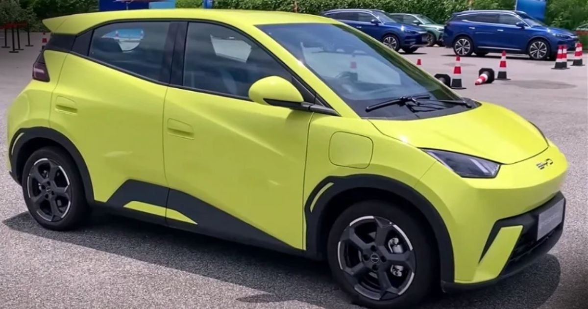 Energia Solar e Veículos Elétricos - Vista lateral de um carro elétrico compacto de cor verde-limão estacionado, com design moderno e aerodinâmico, destacando-se contra um fundo de outros veículos e cones de sinalização.